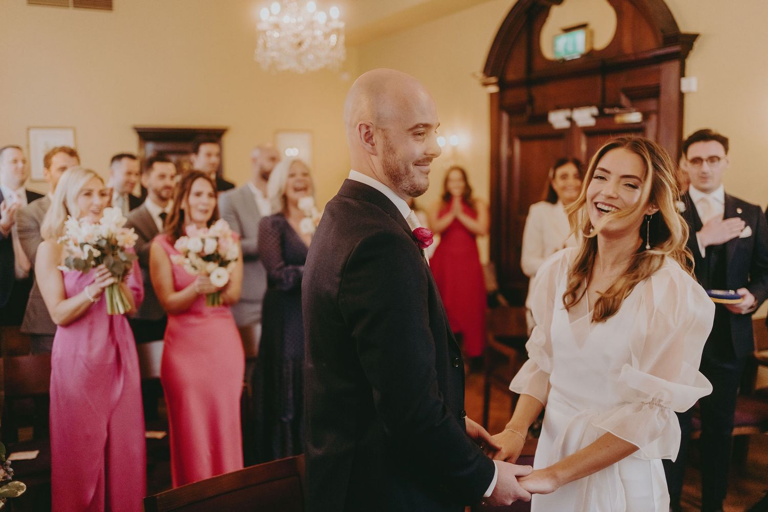A couple exchanges vows during their wedding ceremony with bridesmaids in pink dresses watching in the background.