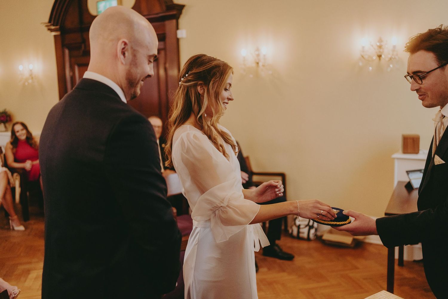A wedding ceremony shows the exchange of rings in a traditional indoor venue with classic architectural details.
