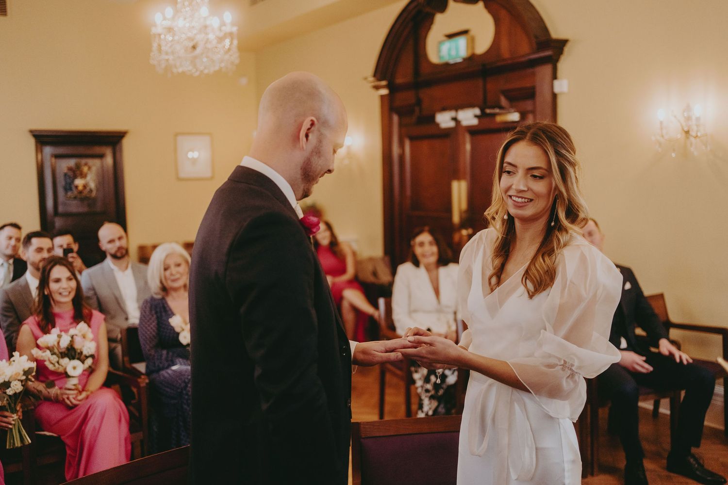 A wedding ceremony unfolds in an intimate indoor setting with guests watching from wooden chairs.
