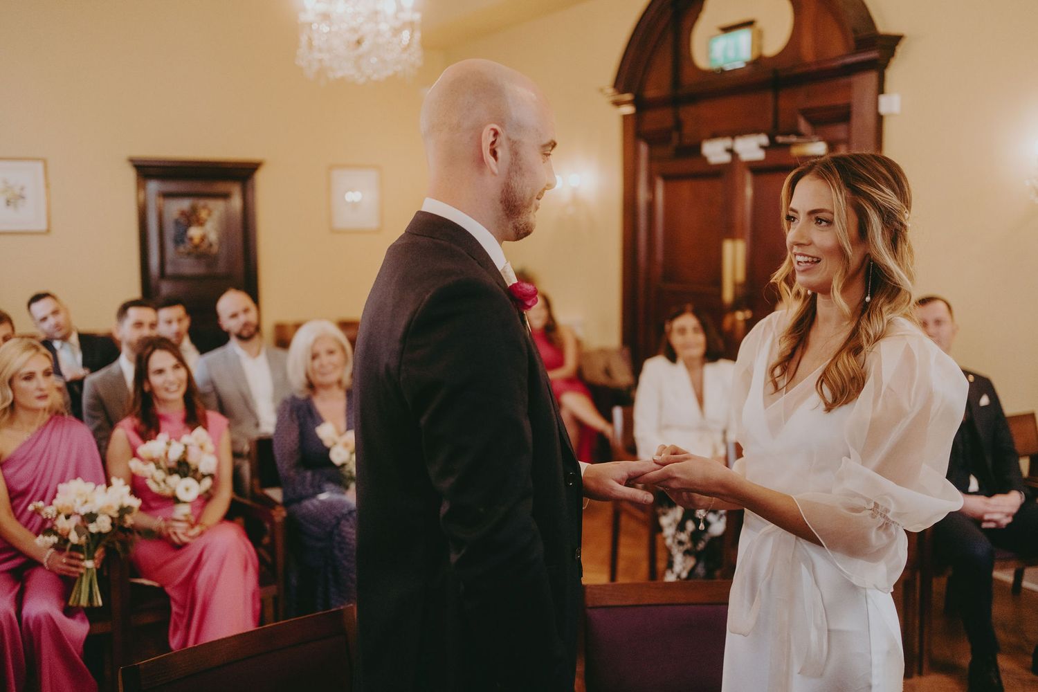 A couple exchanges wedding vows in front of guests seated in a classic ceremony room with chandeliers.