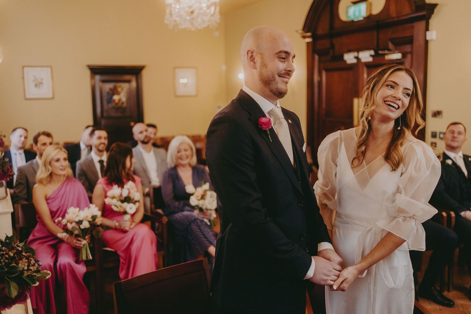 A joyous moment captured during a wedding ceremony with guests and bridesmaids in pink dresses looking on.