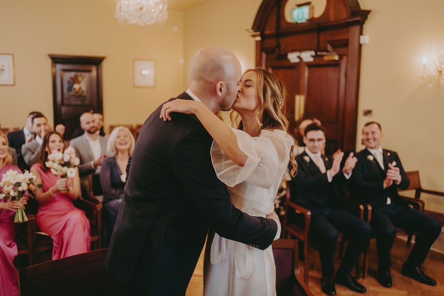 Newlyweds share their first kiss as guests applaud during an intimate indoor ceremony.