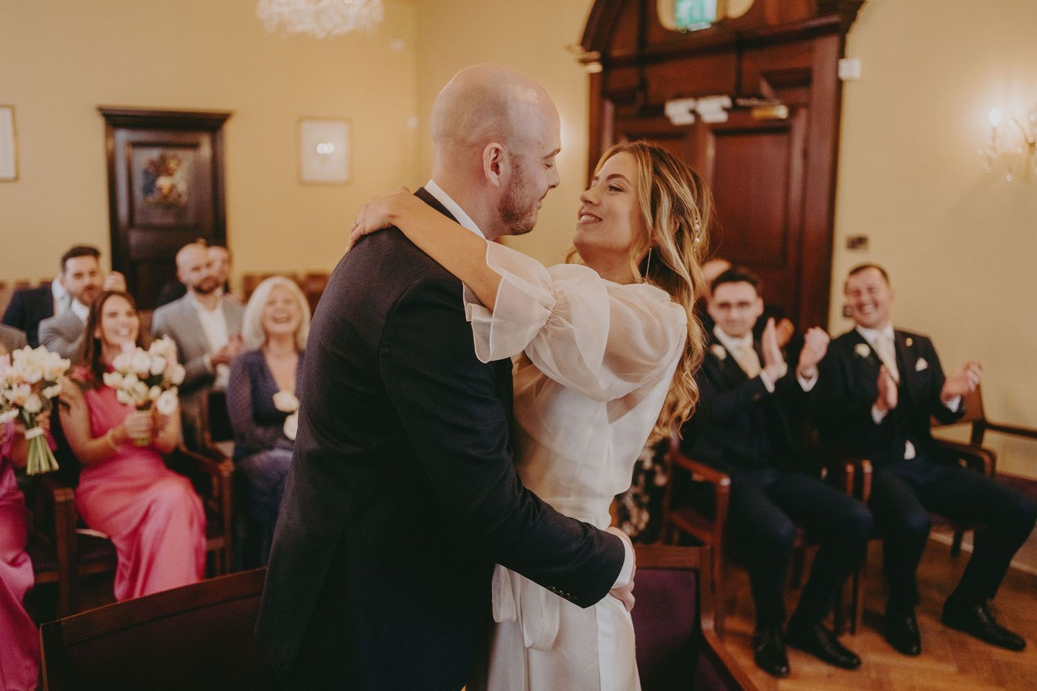 A romantic embrace between newlyweds in a vintage-style reception room with wedding guests looking on.