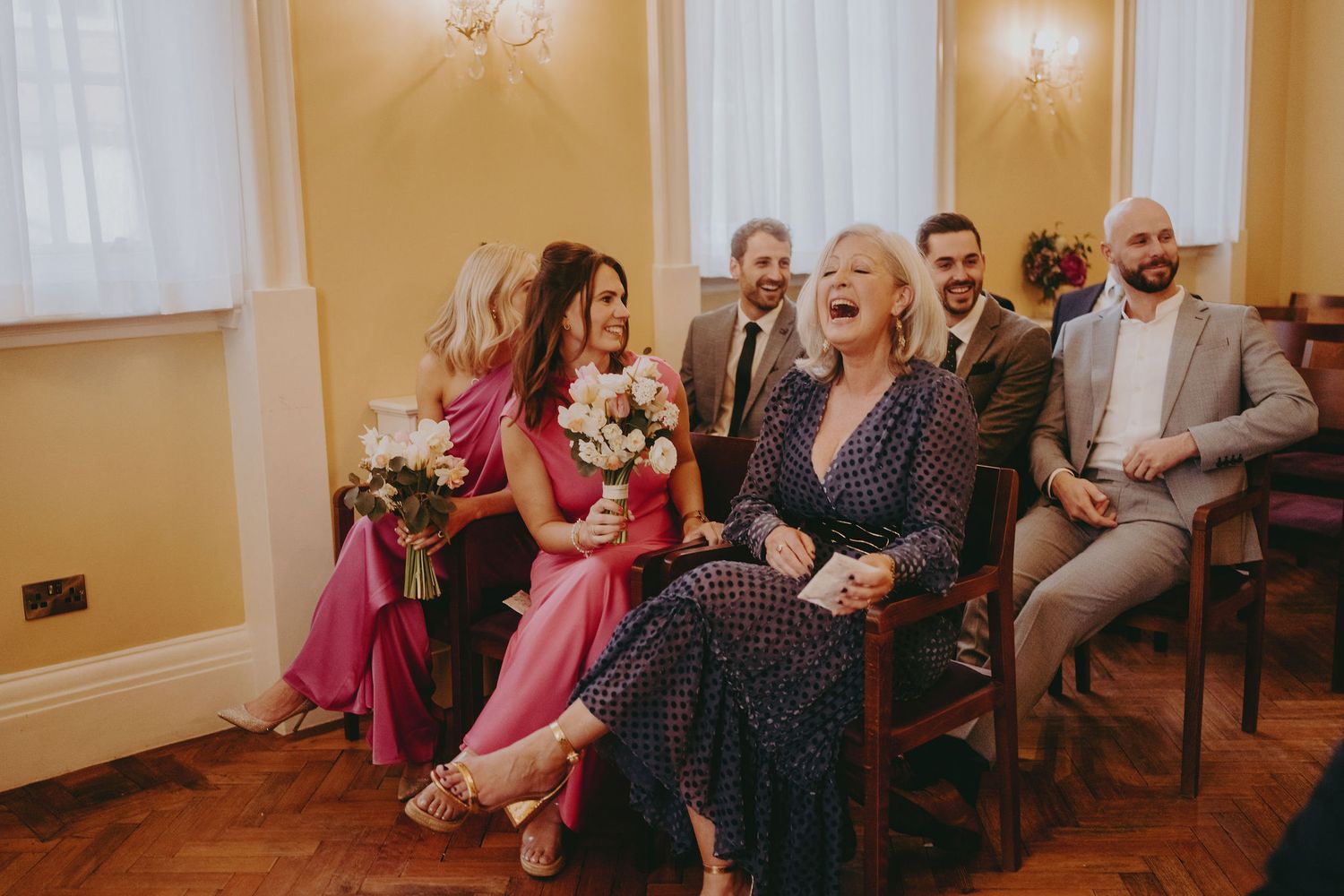 Wedding guests in vibrant attire laugh together during a reception in a warm-lit room.