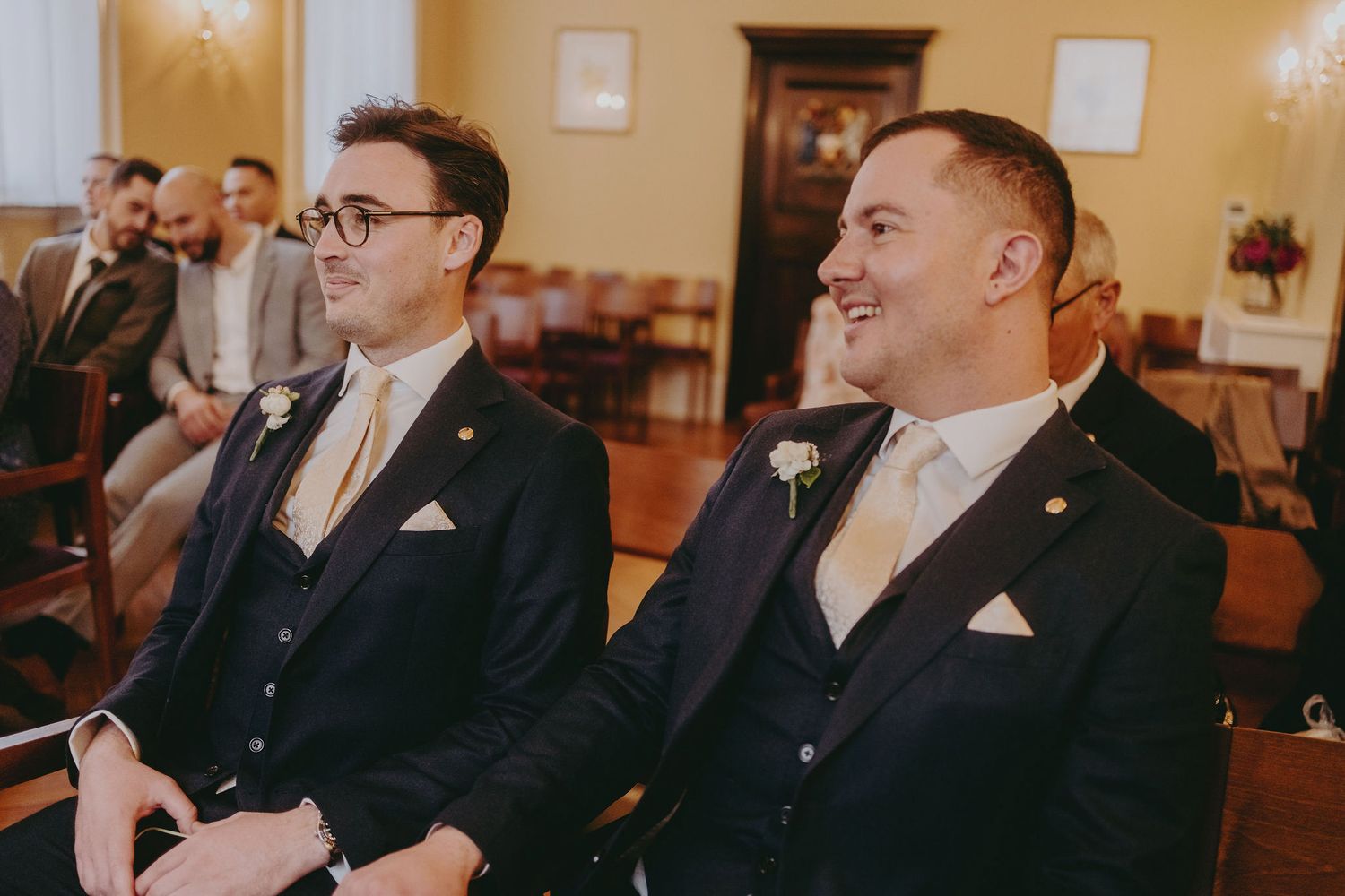 Two groomsmen in matching suits share a laugh during wedding ceremony.