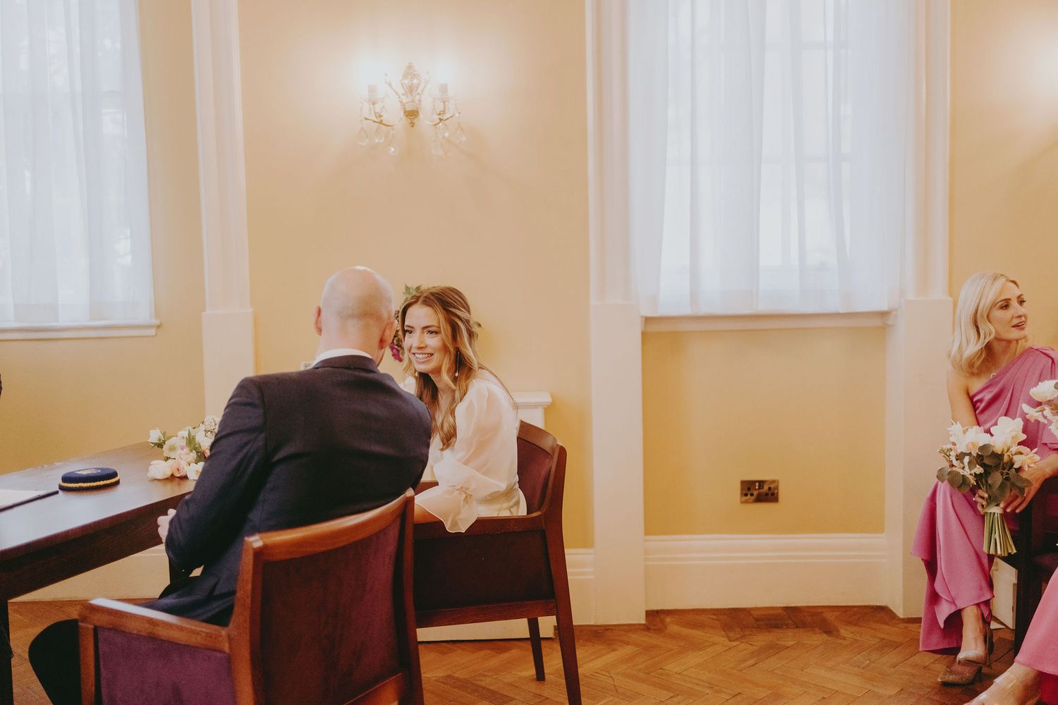 Wedding couple seated at registration table with guests in background of yellow-walled ceremony room.