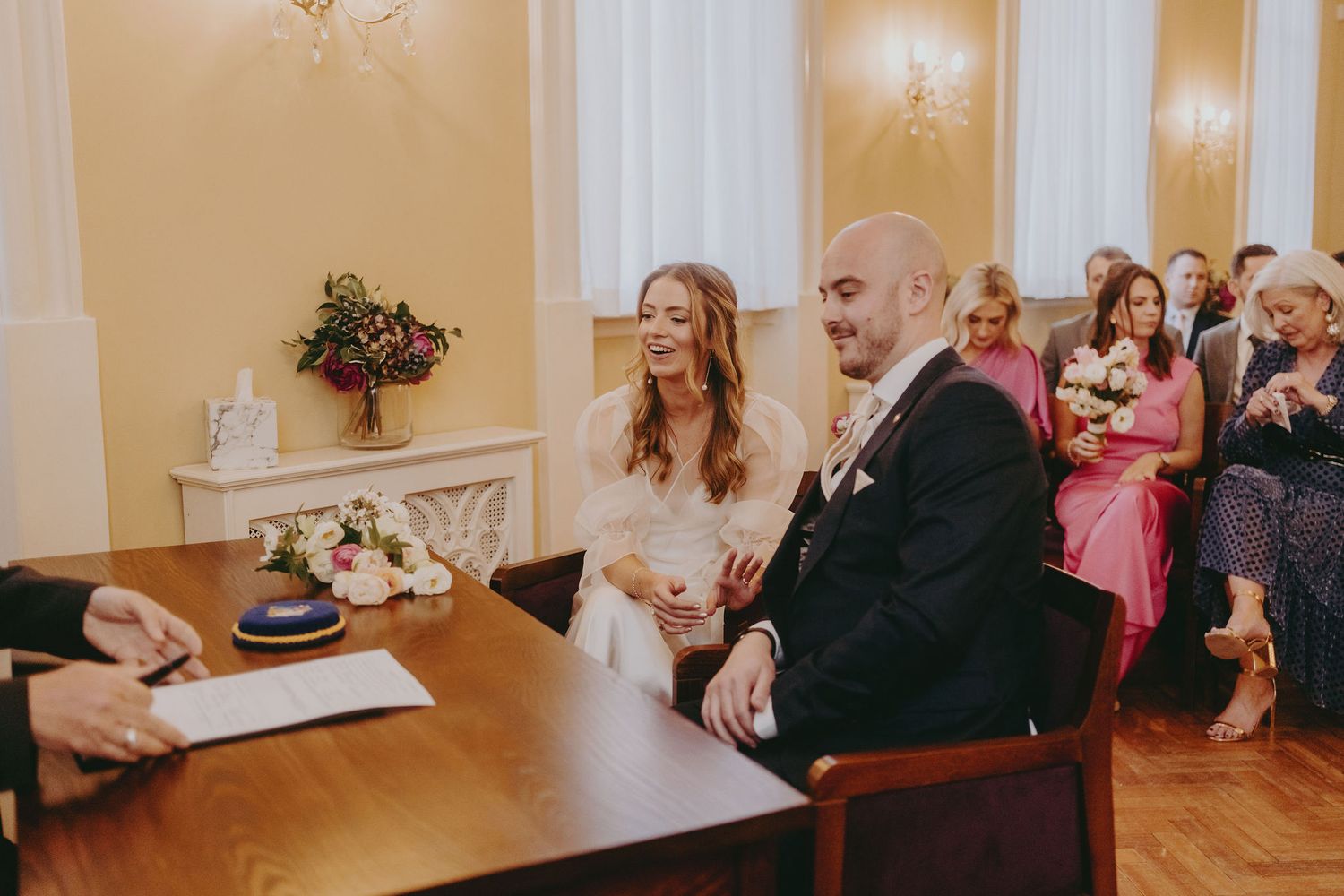 Wedding couple signs marriage documents in an elegant room with guests looking on.
