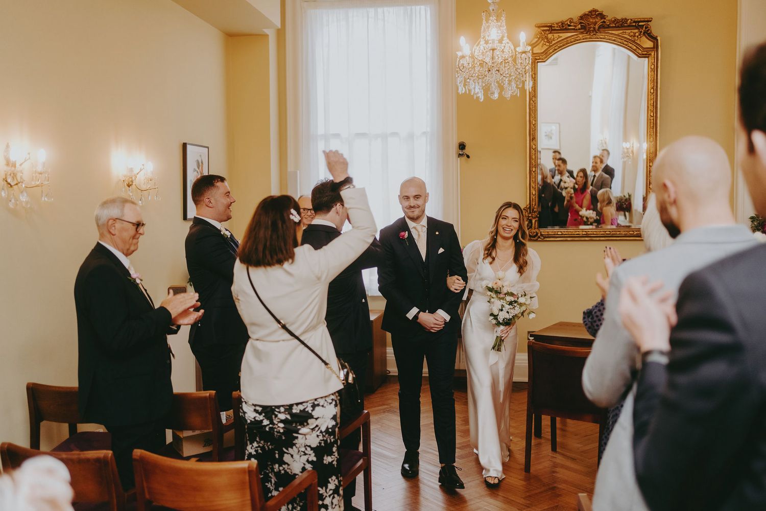Intimate wedding ceremony taking place in a warmly lit room with chandelier and ornate mirror.