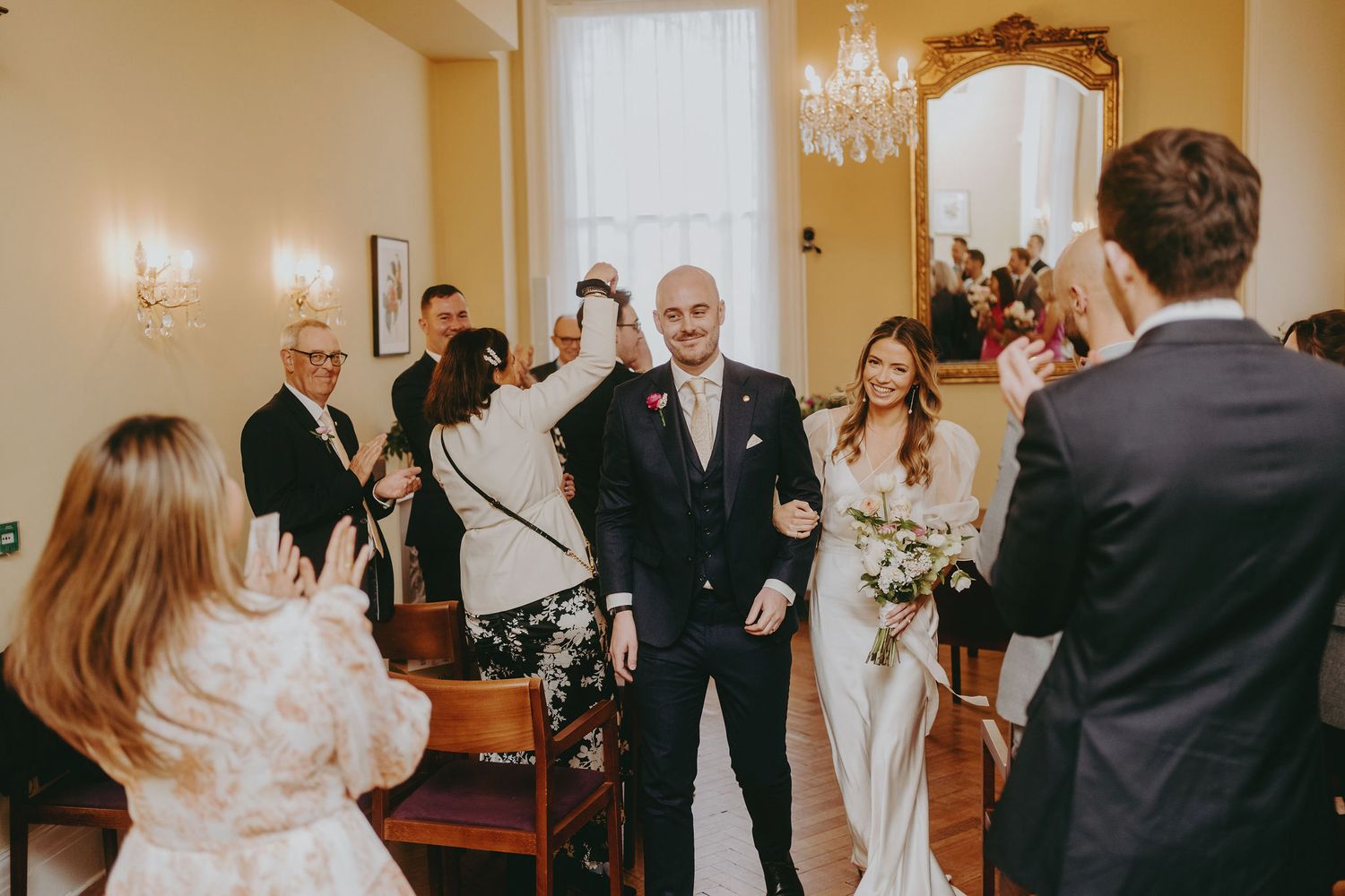 Wedding ceremony in progress in an elegant room with chandelier and decorative mirrors.