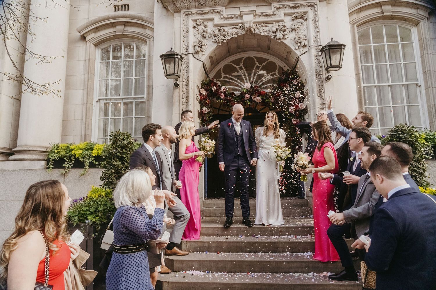 Wedding party poses on grand stone steps of historic building with pink-dressed bridesmaids.