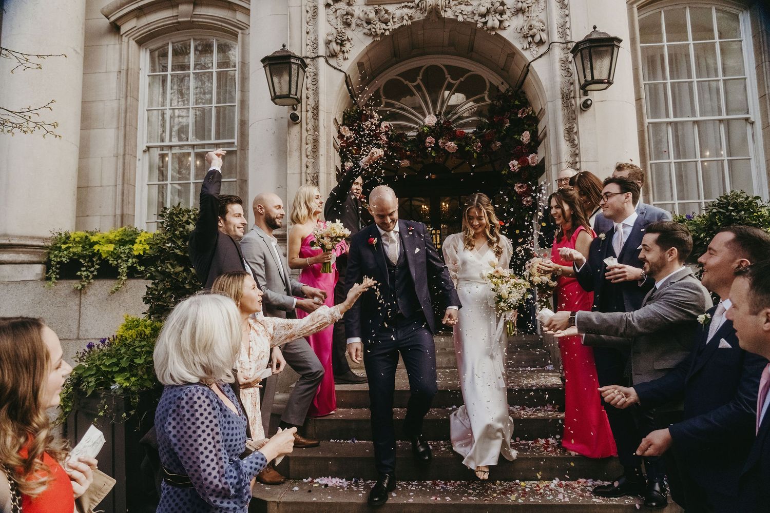 Wedding party celebrates on historic building steps with guests throwing confetti.