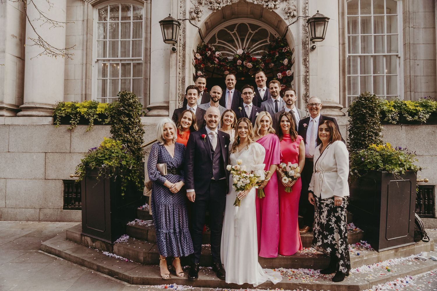 Wedding party gathered on steps of elegant building with ornate architectural details and potted yellow flowers.