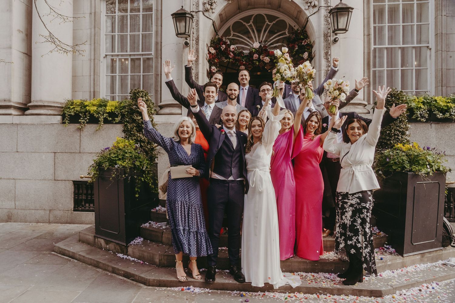 Celebratory wedding group photo with raised arms in front of historic building entrance.