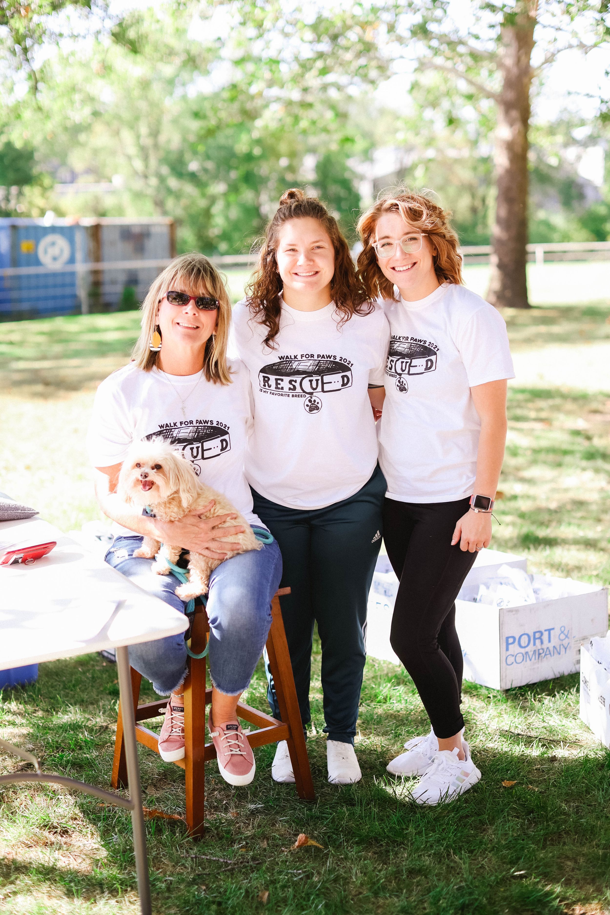 Friends with small dog posing together at outdoor charity event wearing matching shirts.
