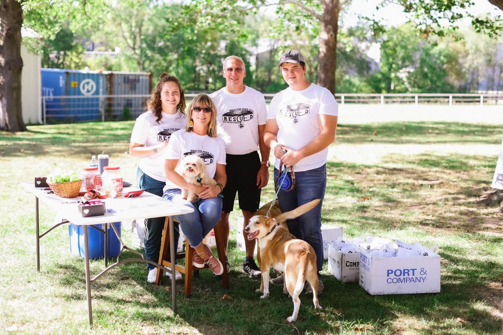 People and dogs gather outdoors at a community event on a sunny day under trees.