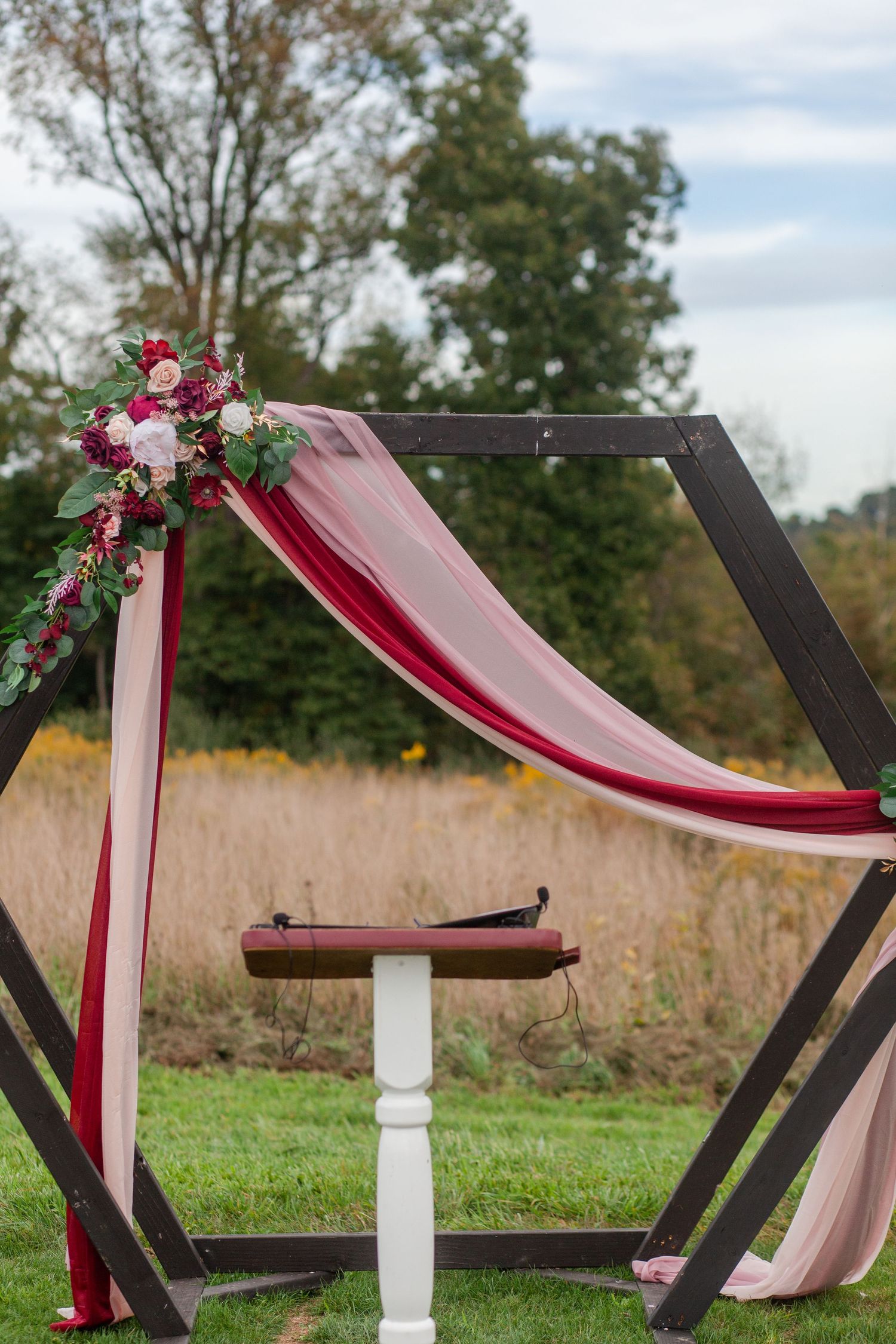Modern black hexagonal wedding arch decorated with burgundy draping and floral arrangements in an outdoor setting.