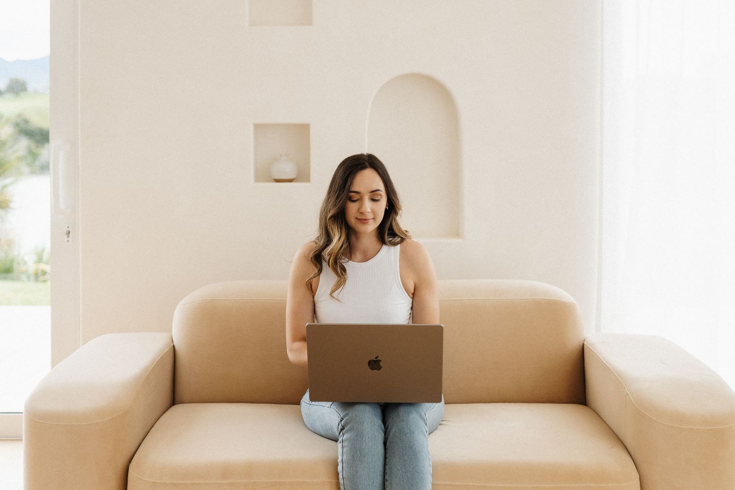 Woman working on laptop while sitting comfortably on couch in minimalist white room.