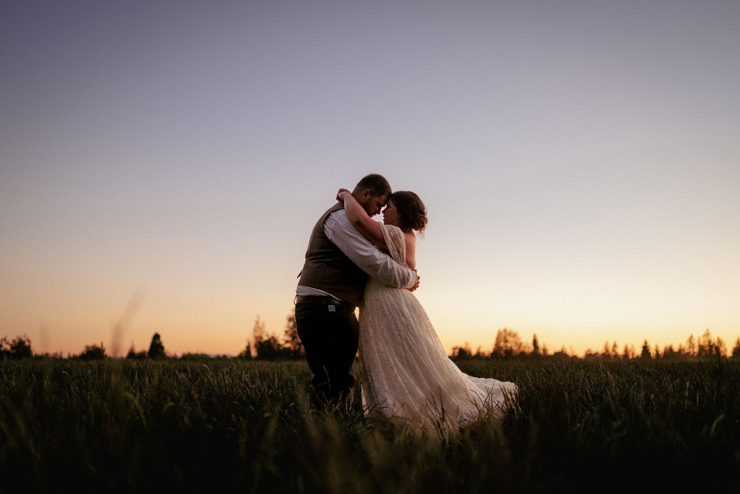 Bride and groom kissing in tall grass during sunset. Intimate golden hour moment.