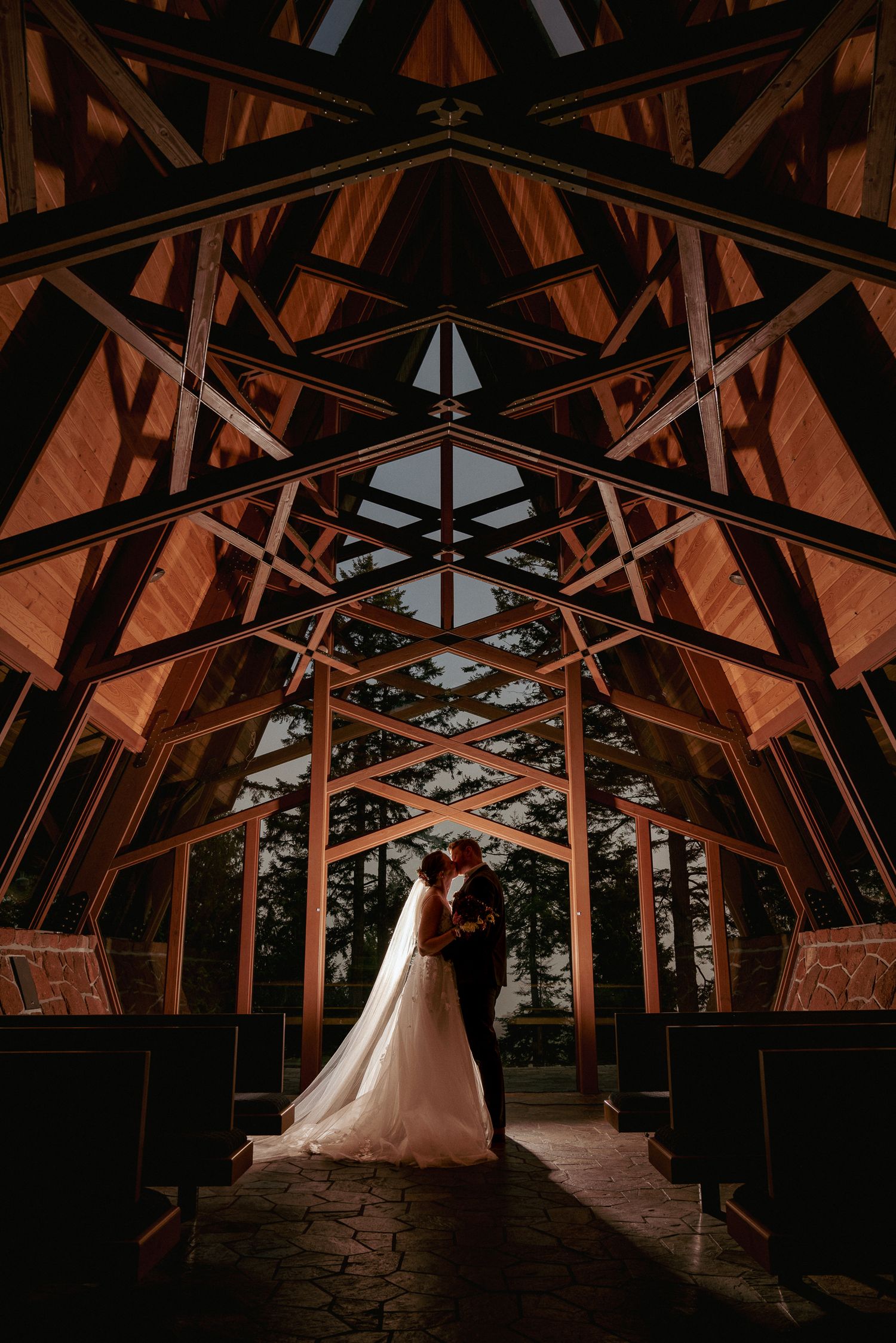 Bride and groom standing at the altar in a stunning modern wooden chapel with dramatic triangular beams and warm light pouring in.