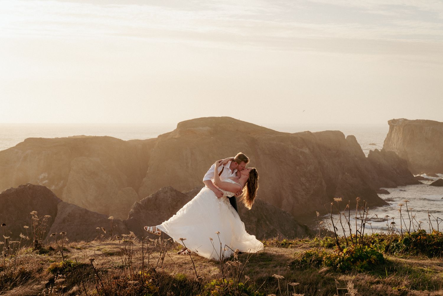 Bride and groom kiss on Oregon Coast cliffs, windswept love story.