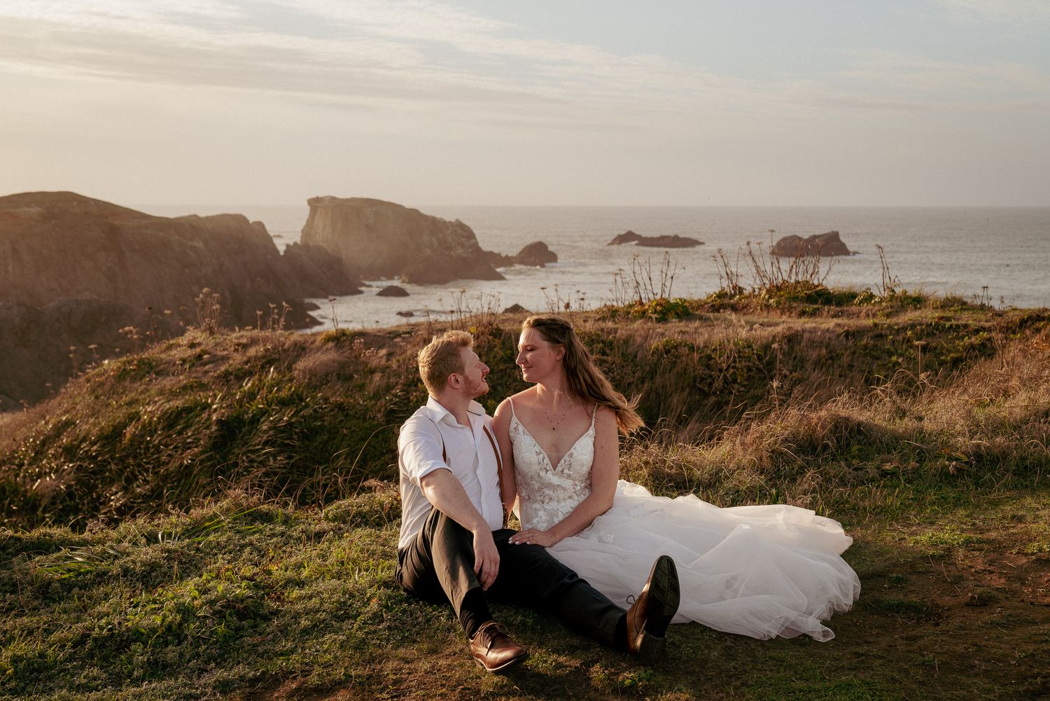 Bride and groom sit atop cliff at sunset, wide-angle shot with Pacific Ocean and rugged cliffs in the background. Cinematic Oregon Coast