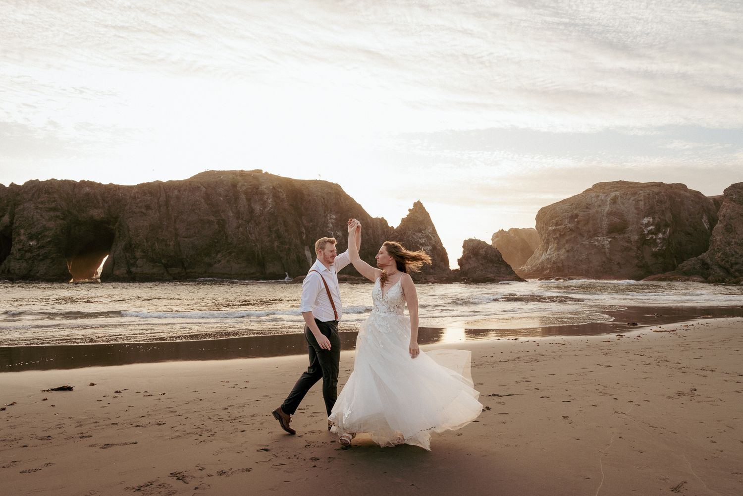 Bride and groom dancing hand-in-hand on Oregon Coast beach at sunset. Romantic coastal wedding moment.
