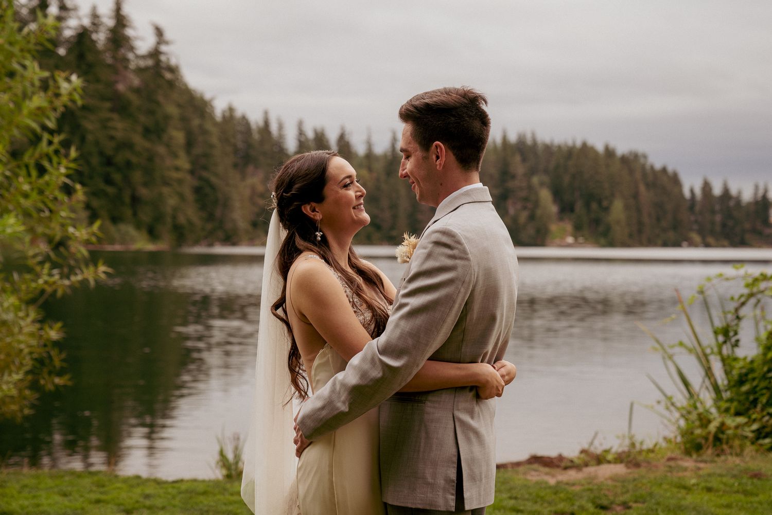 Bride and groom embrace by lake, peaceful Oregon forest elopement.