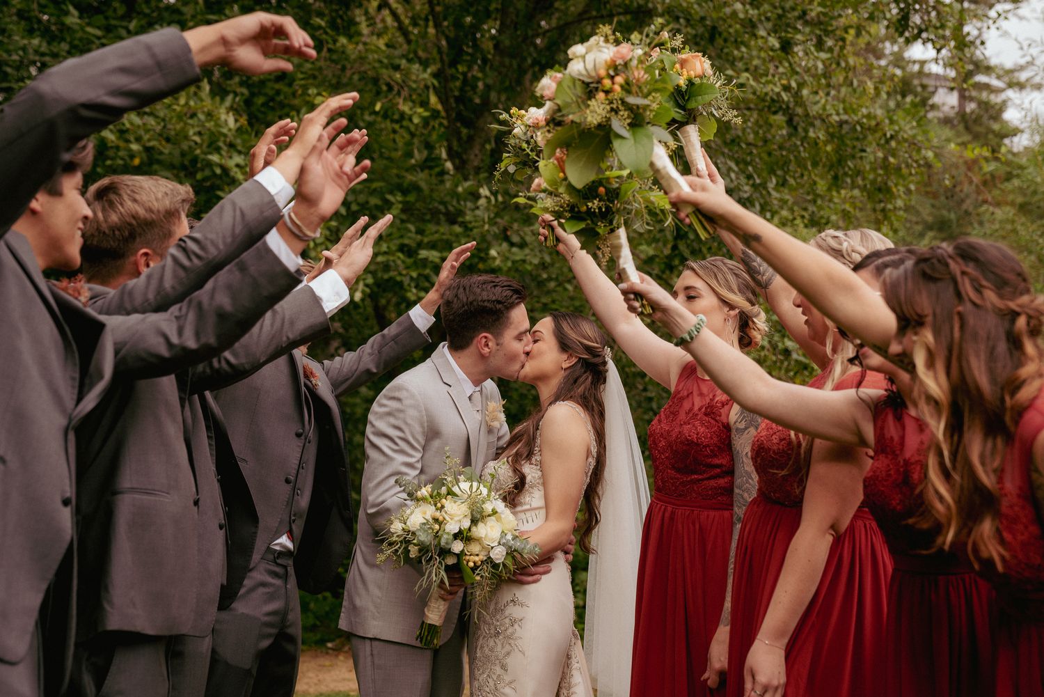 Bride and groom kiss while bridal party cheers, tossing flower bouquets and hands in the air. Joyful wedding celebration moment.