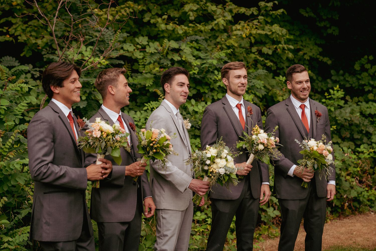 Groomsmen laugh with bouquets in hand, lighthearted forest wedding.