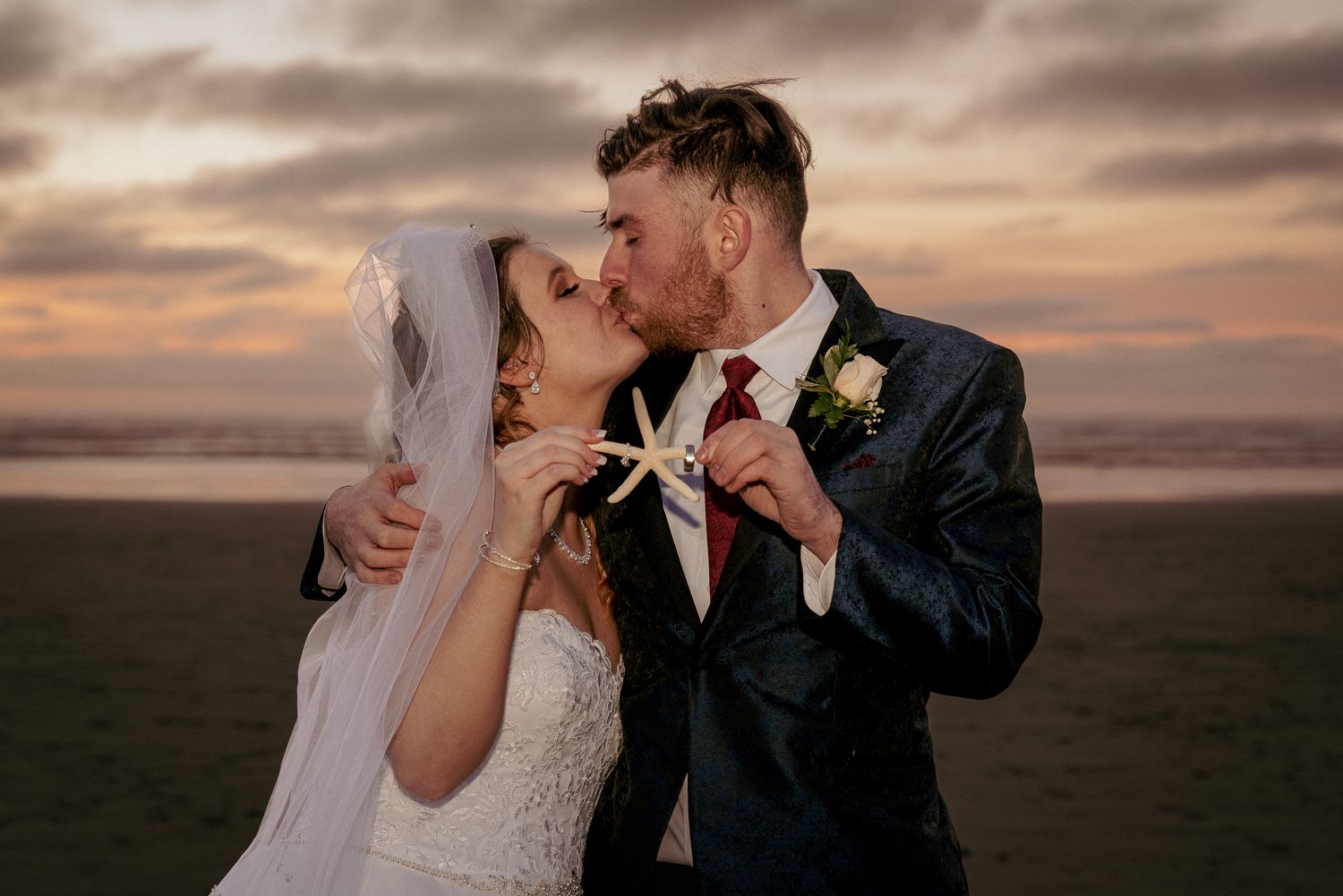 Newlyweds share kiss on Oregon beach, holding a starfish with the wedding rings on it.