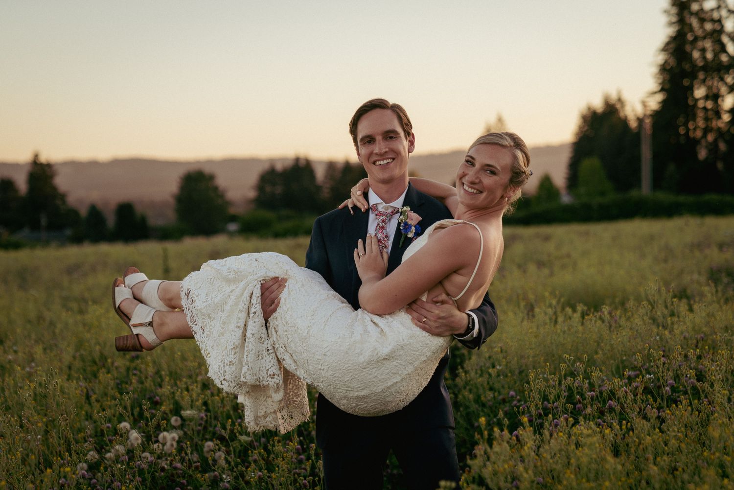 Groom carries bride through meadow, joyful PNW wedding celebration.