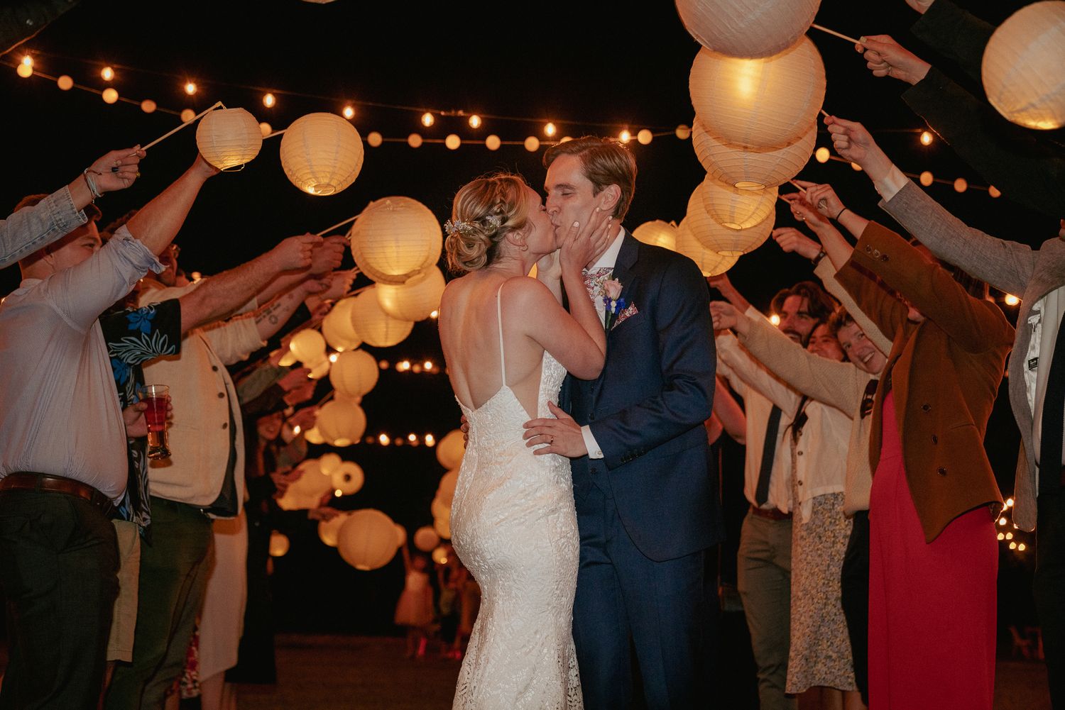 Bride and groom kiss during sparkler send-off under glowing lanterns.