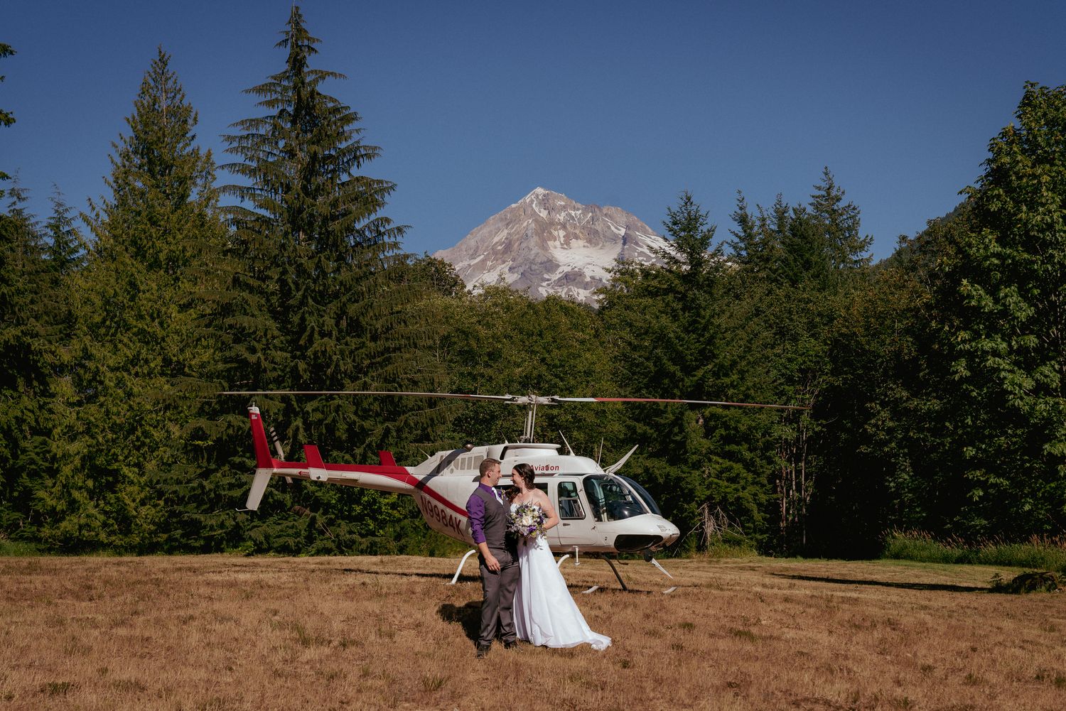 Bride and groom kiss in front of a helicopter with Mount Hood in the background during an adventurous wedding in Oregon.