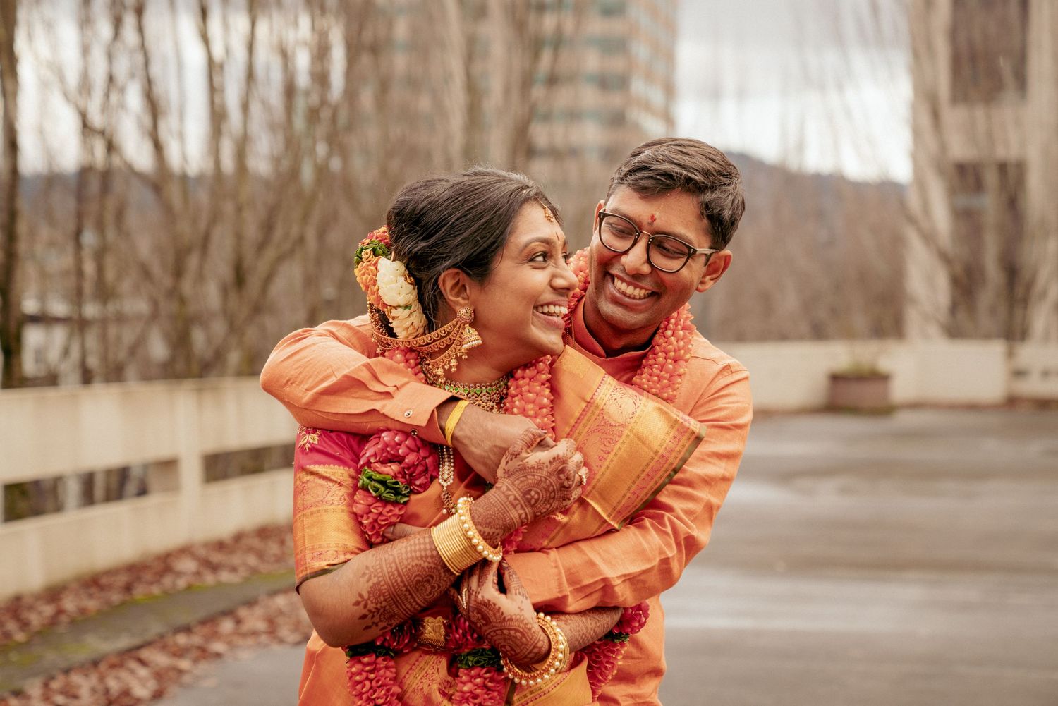 Bride and groom in Indian wedding attire embracing joyfully in urban outdoor setting.