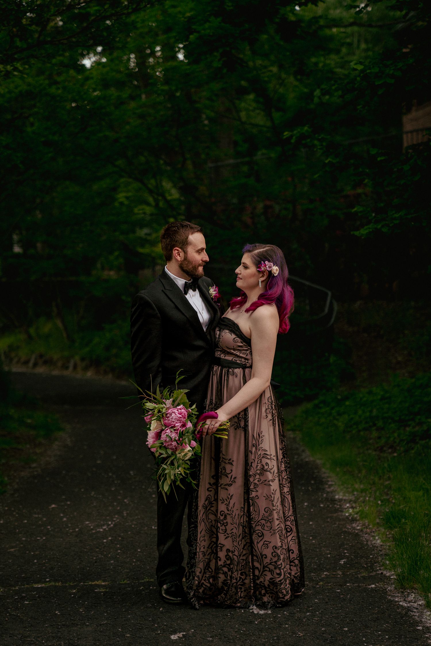 Bride with purple hair and groom smiling at each other outdoors. Unique and joyful wedding portrait.