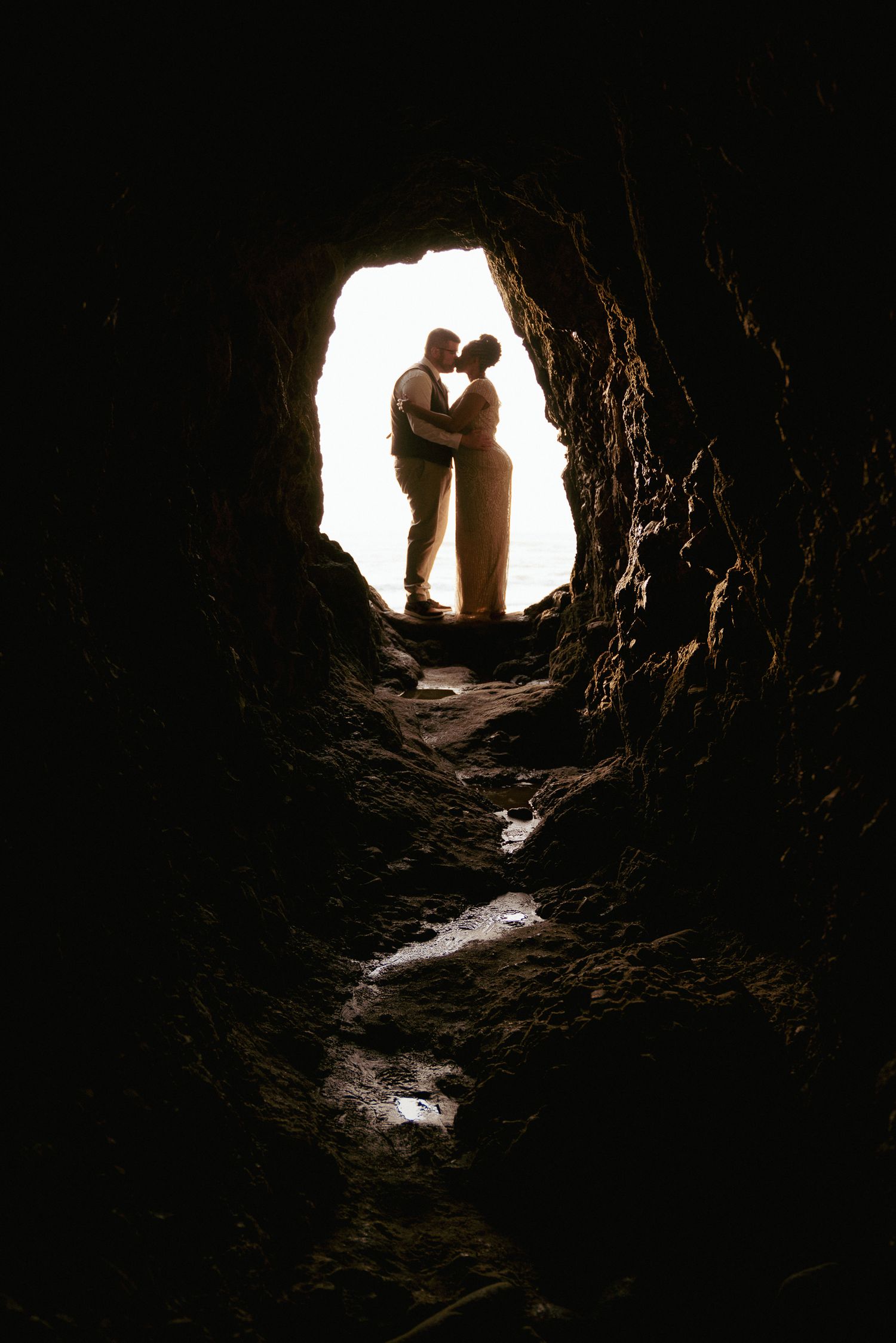 Silhouetted couple sharing an intimate moment inside a dark cave, with bright daylight behind them creating a heart-shaped frame.