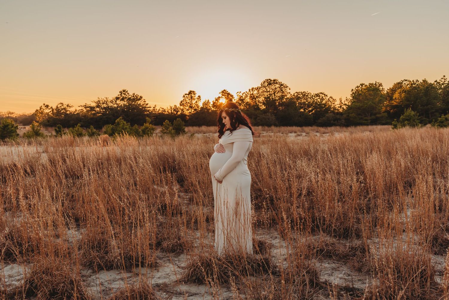 Ethereal maternity portrait in a golden field at sunset with flowing white dress.