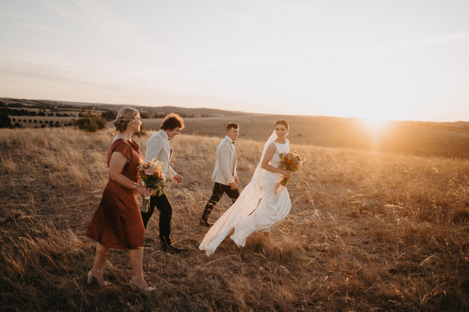 Wedding party walks through a golden field at sunset with flowing dresses and attire catching the warm light.