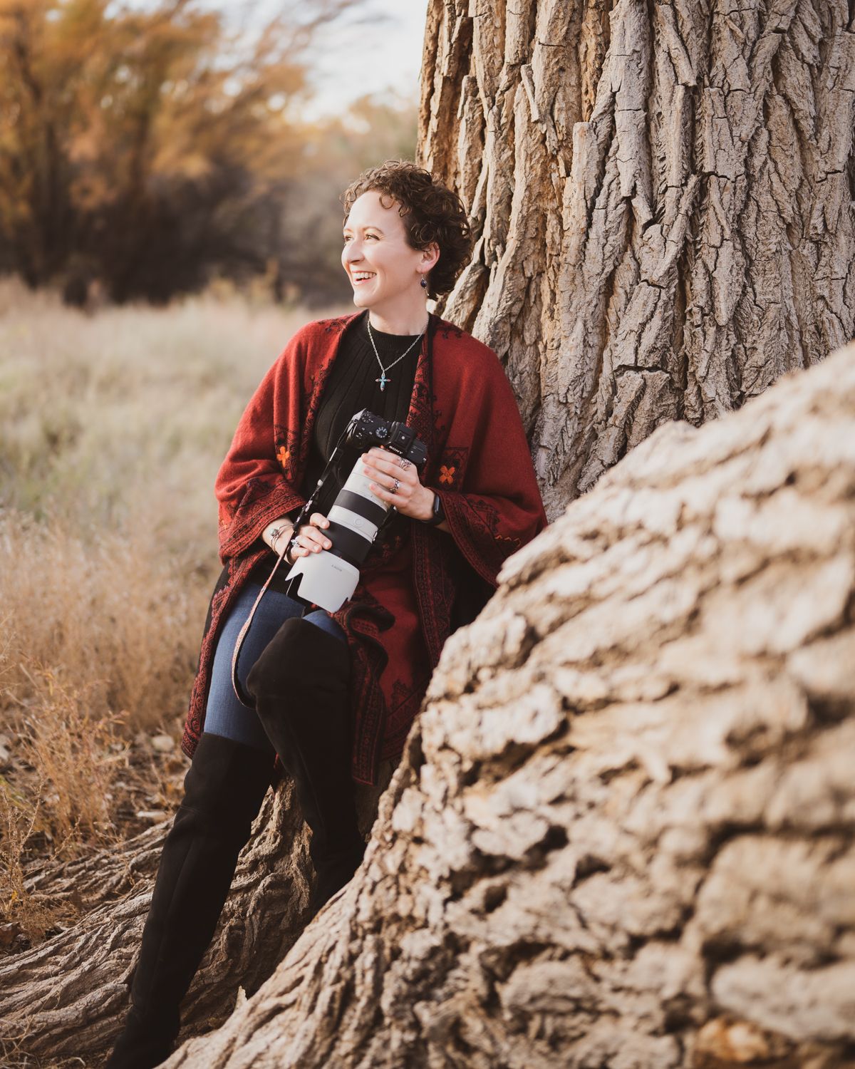 Person in red sweater sits against large tree trunk during fall photoshoot.