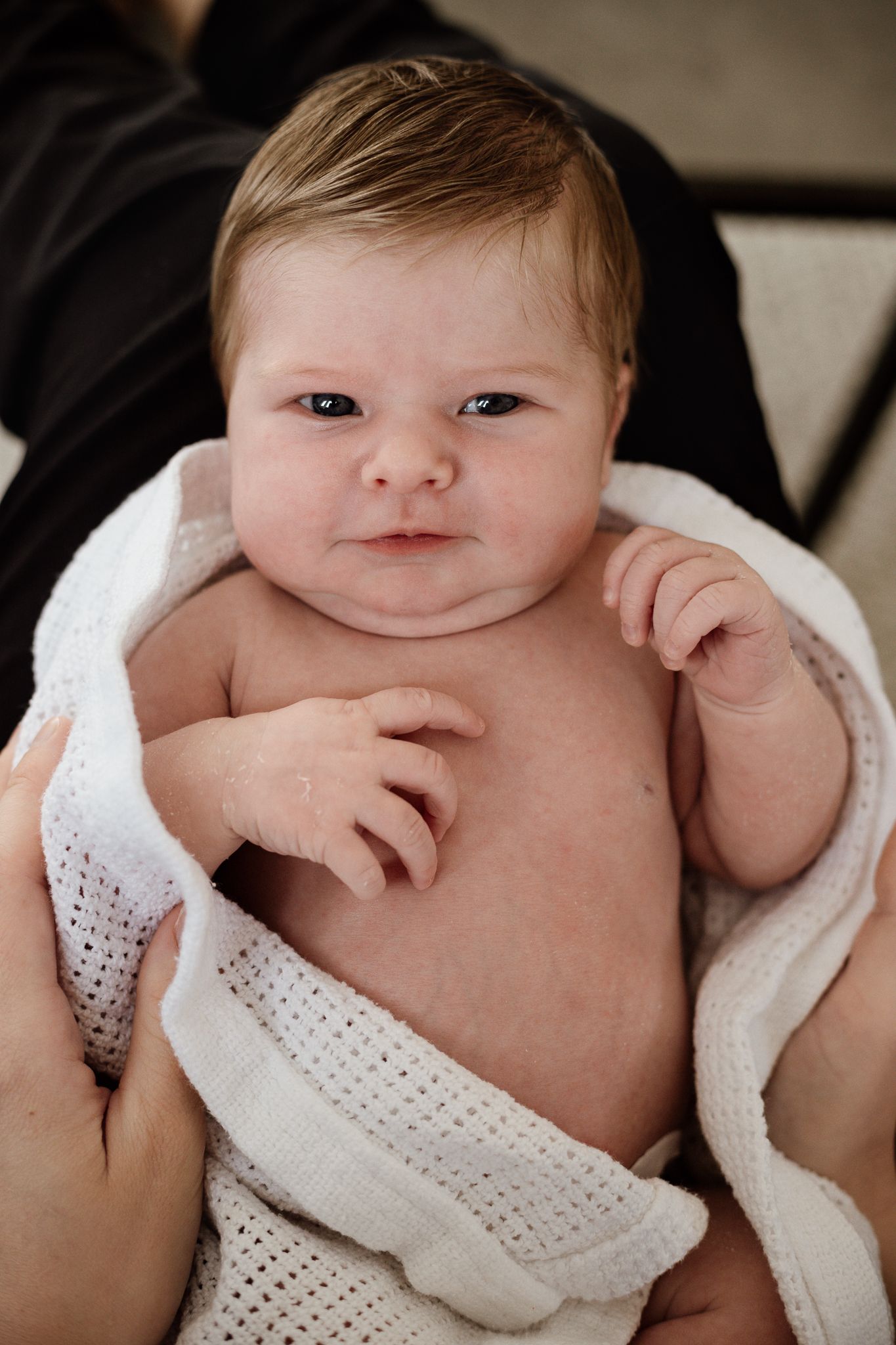 A baby wrapped in a white knit blanket smiling at the camera in a close-up portrait.