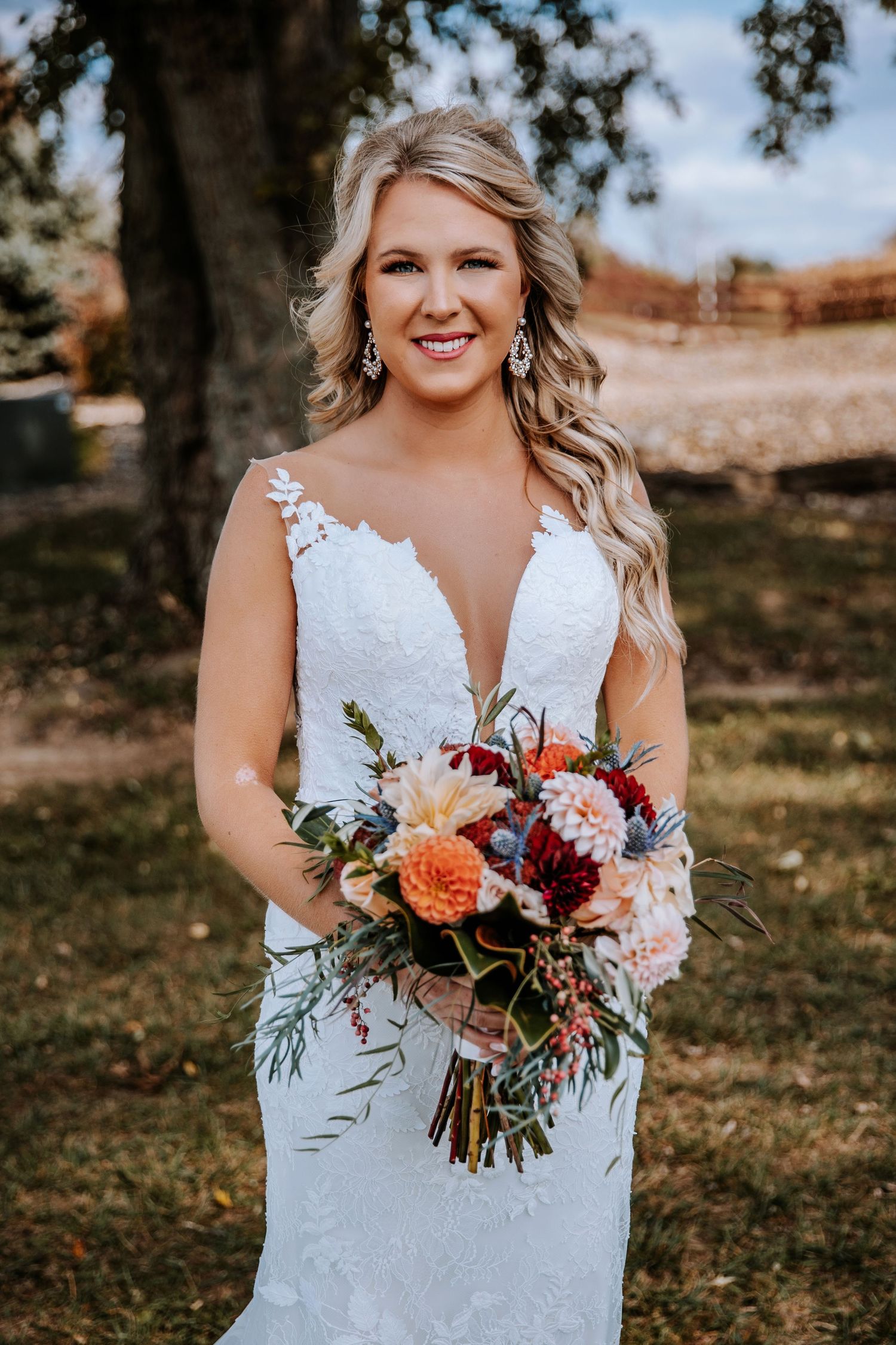 A bride in a white lace gown holds a vibrant autumn bouquet with orange and burgundy flowers in an outdoor setting.