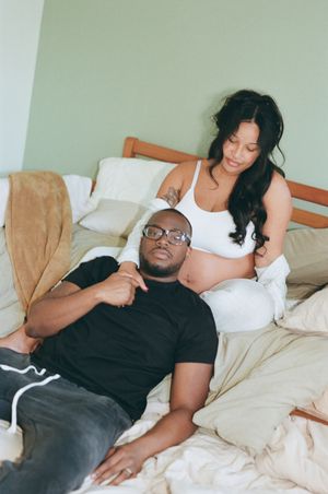 A couple relaxes on a bed together, the woman wearing white is pregnant while her partner wears a black t-shirt.