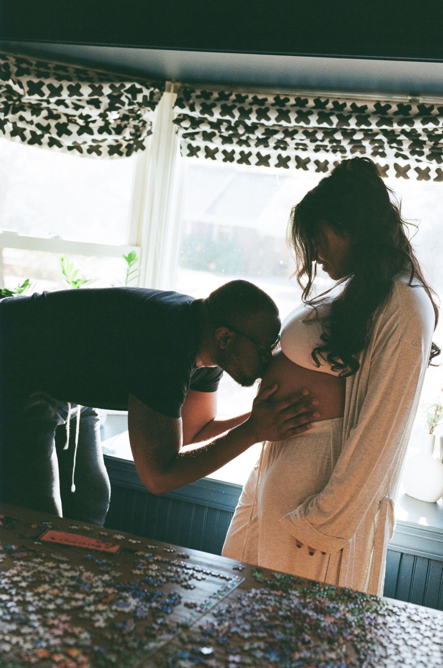 An intimate pregnancy photoshoot shows a tender moment between partners near a window with patterned curtains.