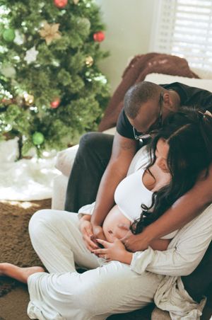 A couple sits intimately near a decorated Christmas tree in their home, wearing white and black clothing.