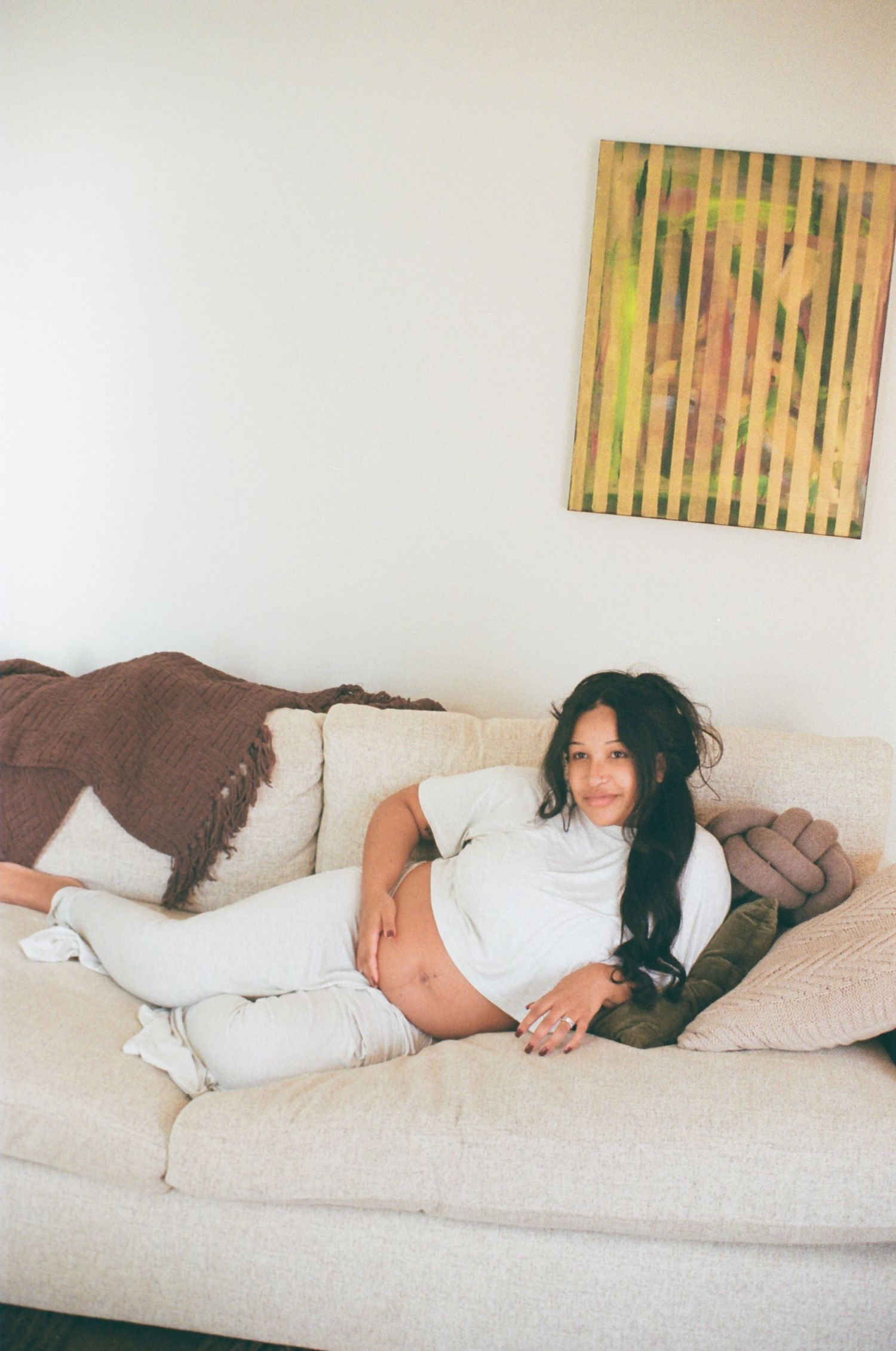 Lounging on a cream couch with a striped artwork on the wall, wearing all white casual attire in a bright interior space.