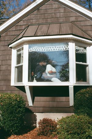 A bay window with ornate white trim on a brown shingled house shows a silhouette sitting inside during daylight hours.