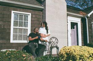 Two people sit together on a porch with white trim and yellow flowers blooming in the garden.