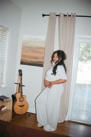 Someone in a flowy white dress stands near a window at home with an acoustic guitar propped against the wall.