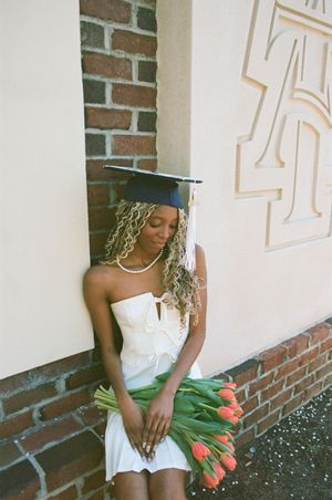 A graduate in a white dress and cap poses with orange tulips against a brick wall on graduation day.