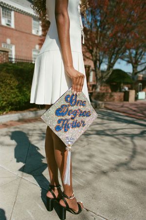 Graduate in white dress holding decorated cap with 'You All See Me Later' text standing on sidewalk near brick building.